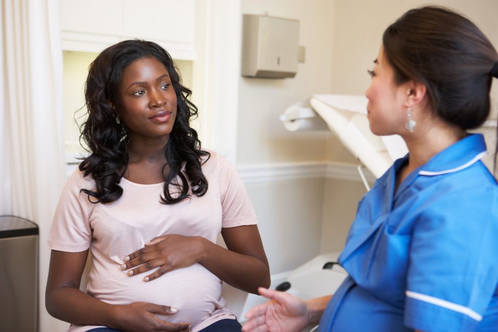 A black pregnant woman talking to an Asian female nurse in a medical office.